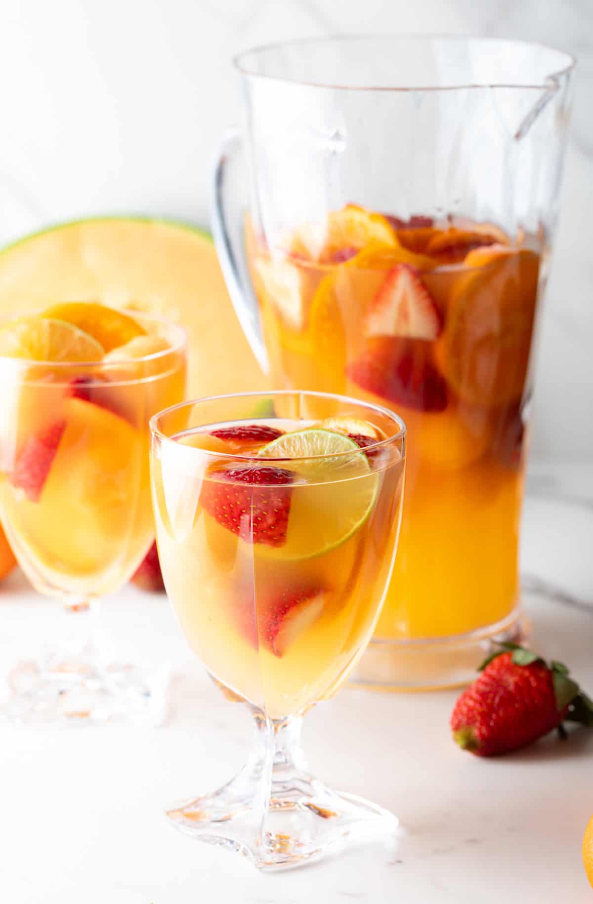 A pitcher and a wine glass filled with a white wine sangria recipe with fresh fruit floating in each.