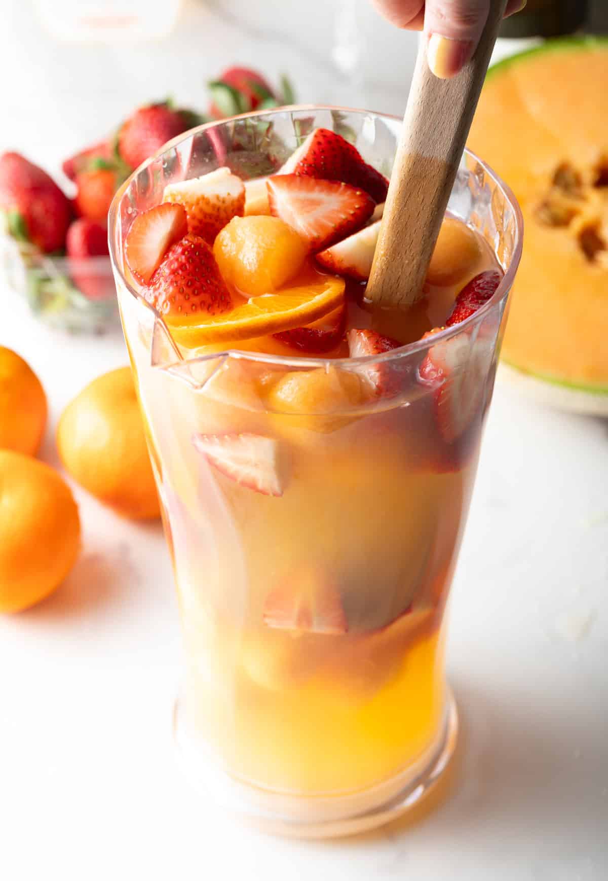 Fresh fruit is being stirred into a pitcher full of white wine sangria.