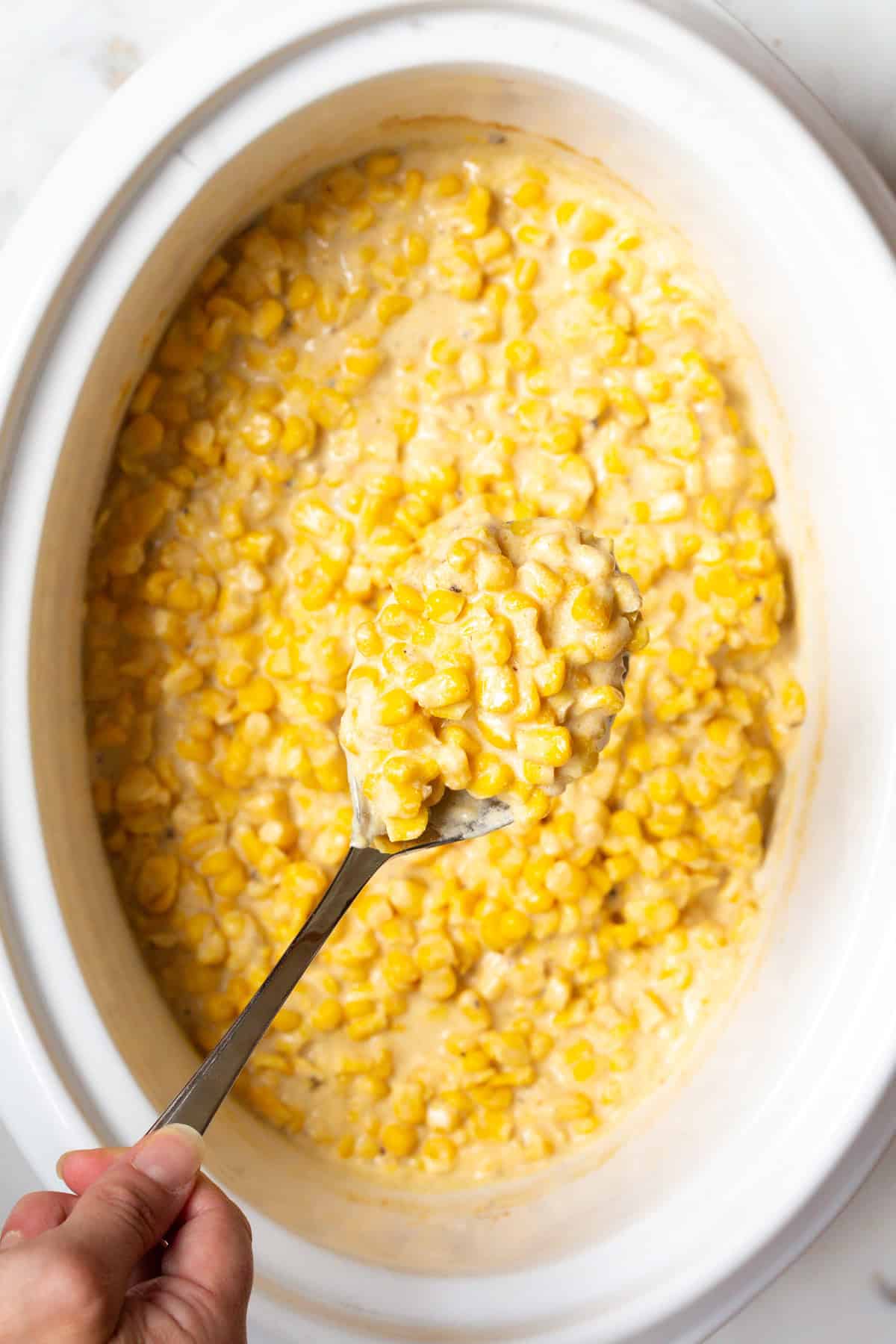 A metal spoon is holding a scoop of crock pot creamed corn above a crock full of the corn recipe.