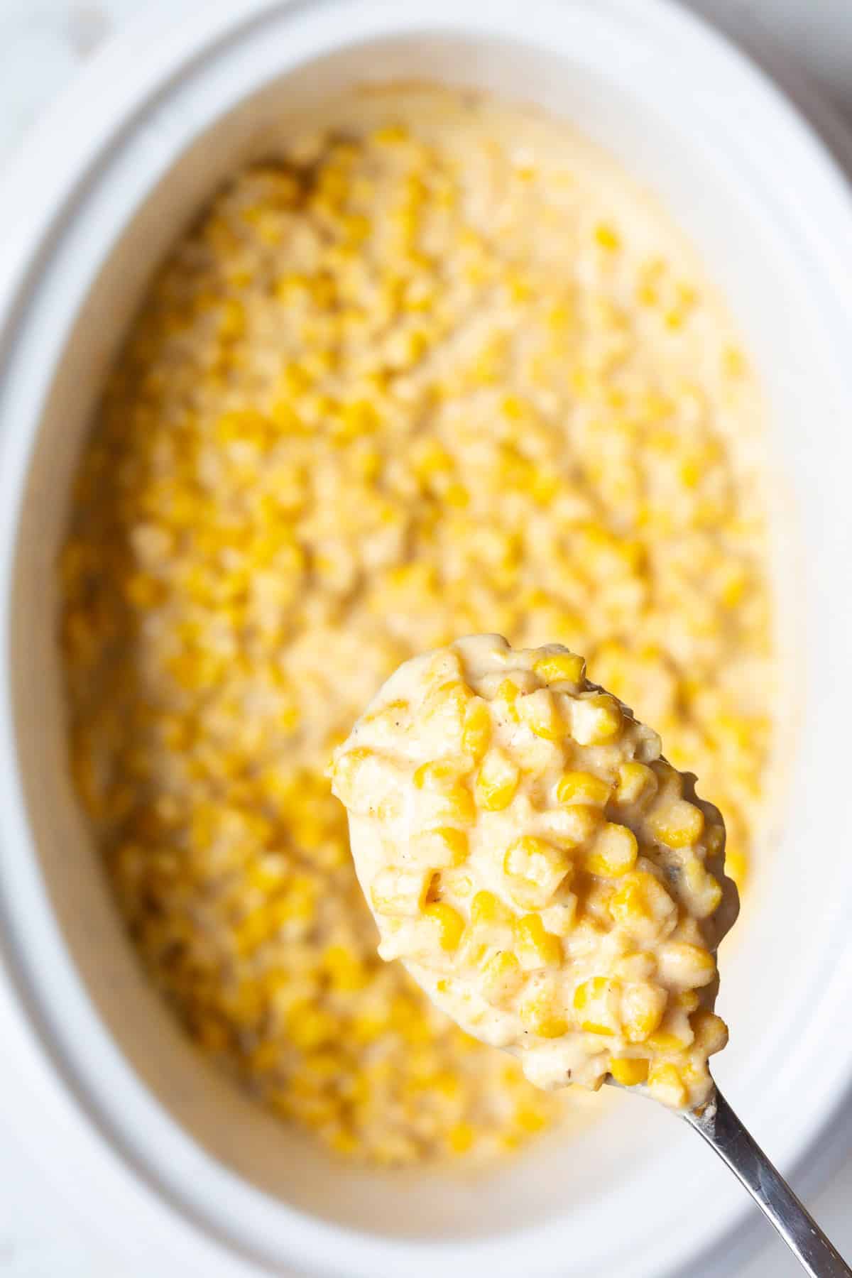A metal spoon is holding a scoop of easy crockpot creamed corn to the camera, above a crock full of the corn recipe.