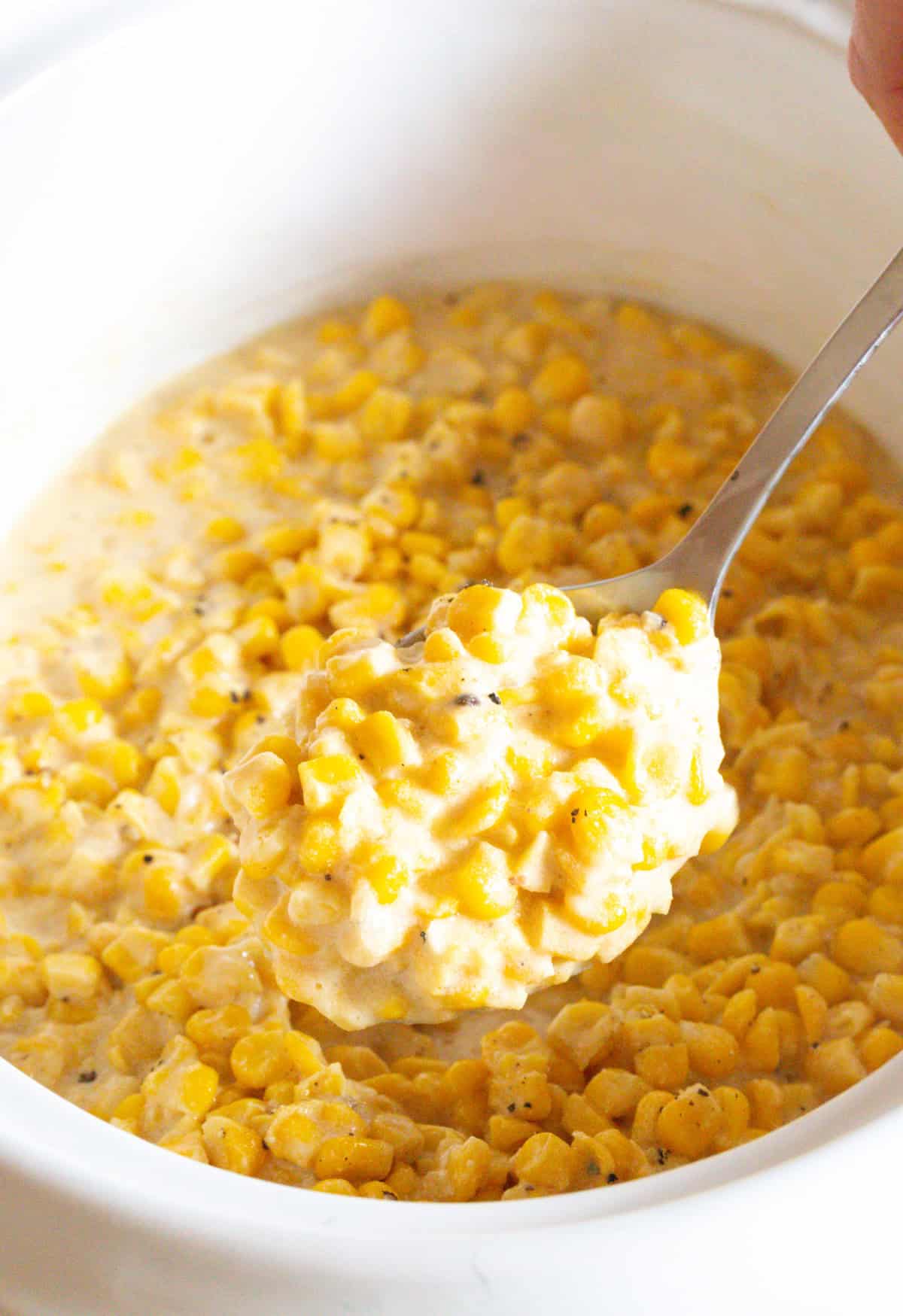 A metal spoon is holding a scoop of easy slow cooker creamed corn to the camera, above a crock full of the corn recipe.