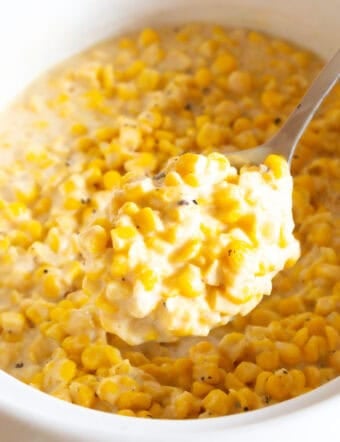 A metal spoon is holding a scoop of crockpot creamed corn to the camera, above a crock full of the corn recipe.