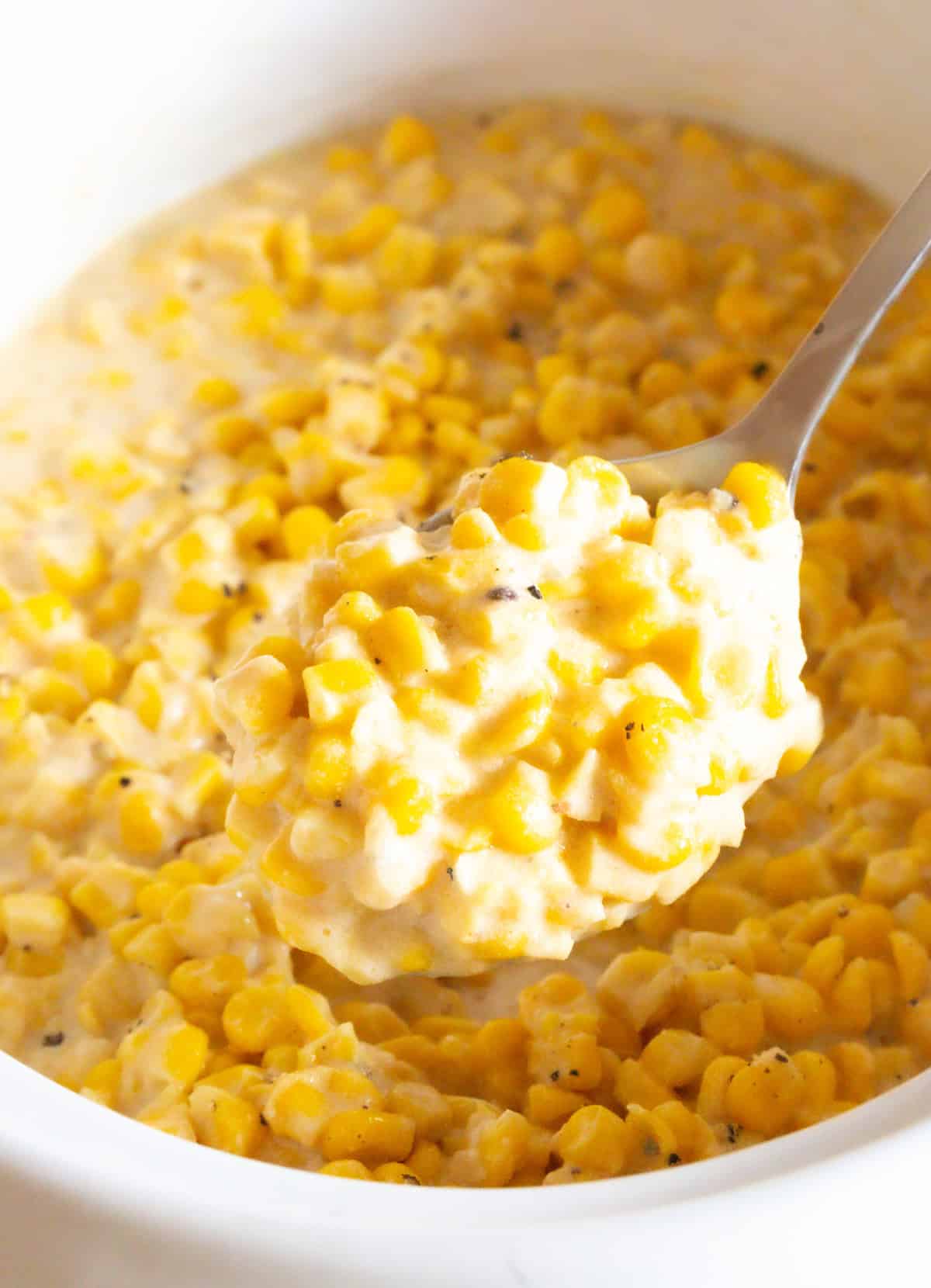 A metal spoon is holding a scoop of crockpot creamed corn to the camera, above a crock full of the corn recipe.
