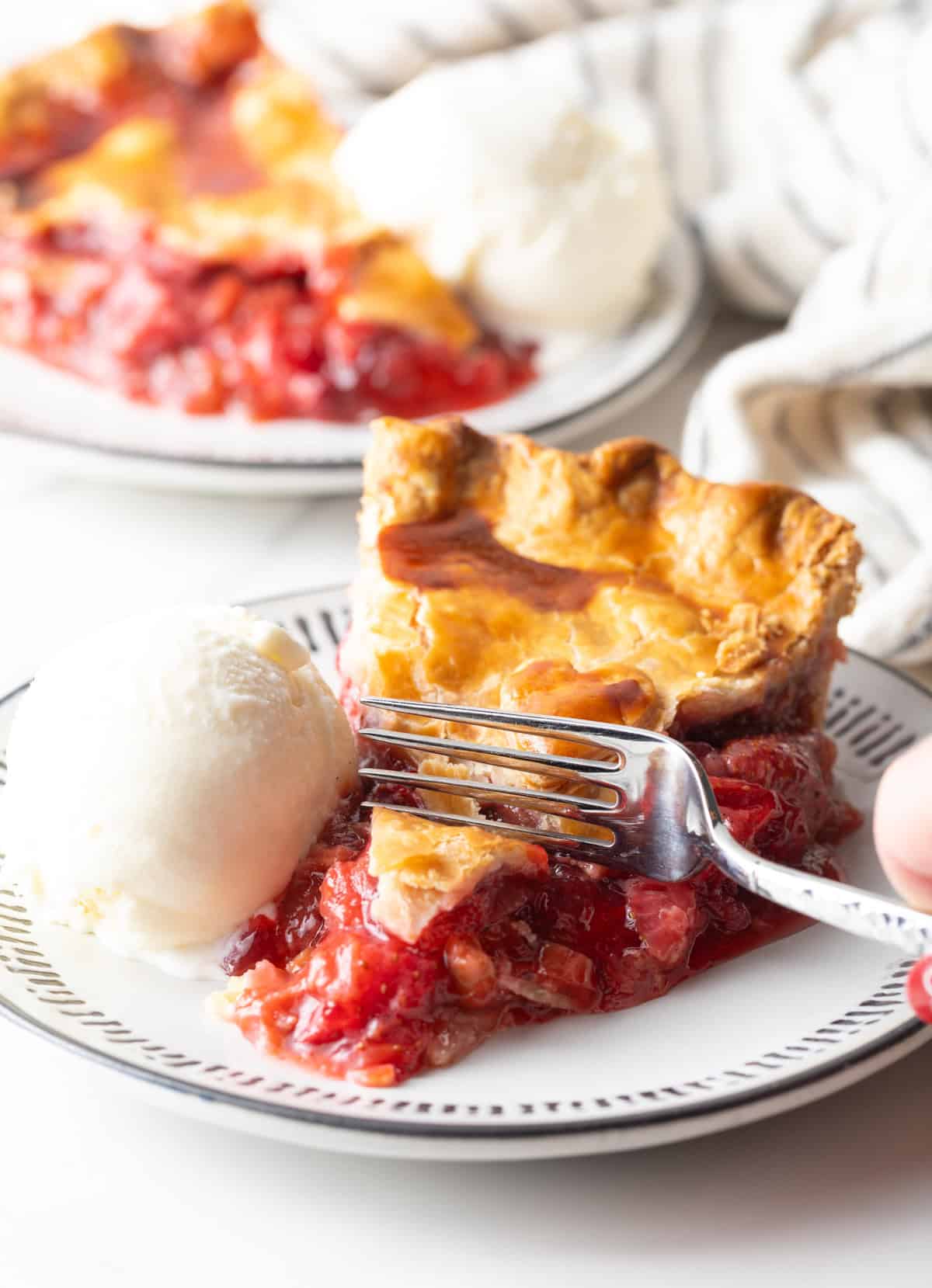 A slice of homemade rhubarb strawberry pie is sitting on a plate with ice cream, and a fork is taking a bite out of the pie.