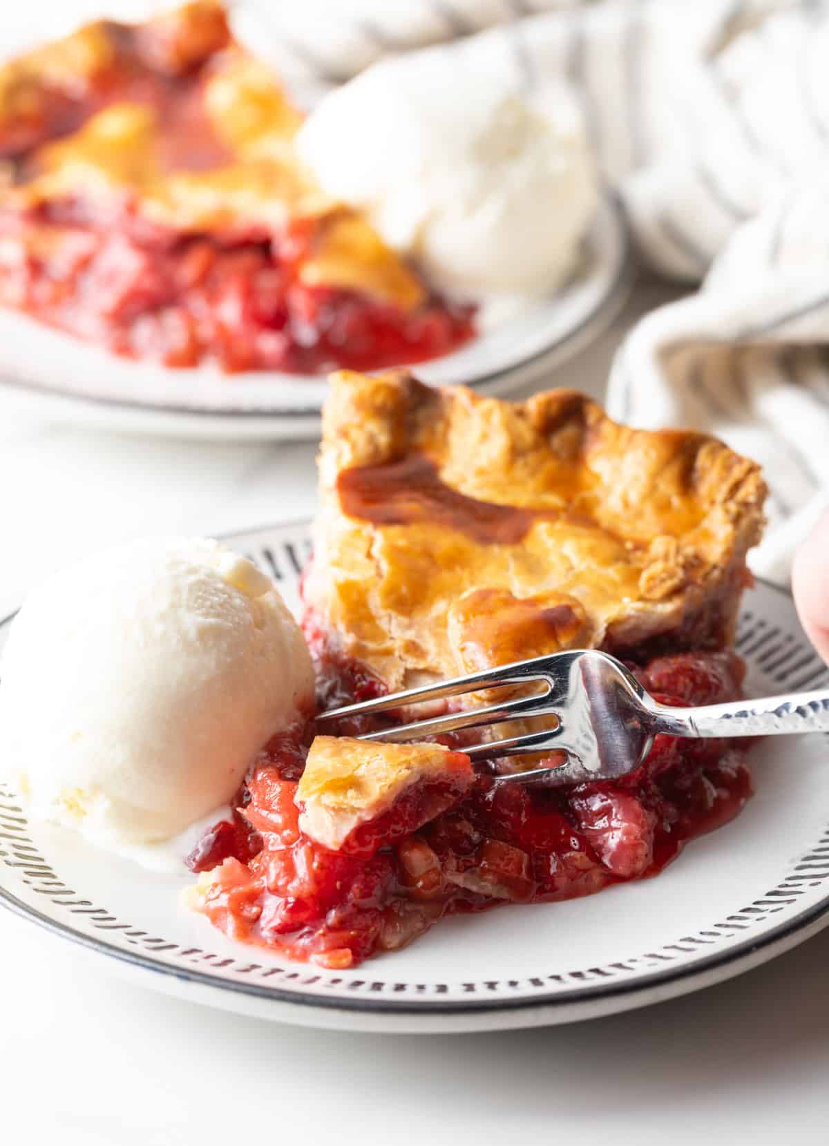 A slice of homemade strawberry rhubarb pie sitting on a plate with ice cream, and a fork is taking a bite out of the pie.