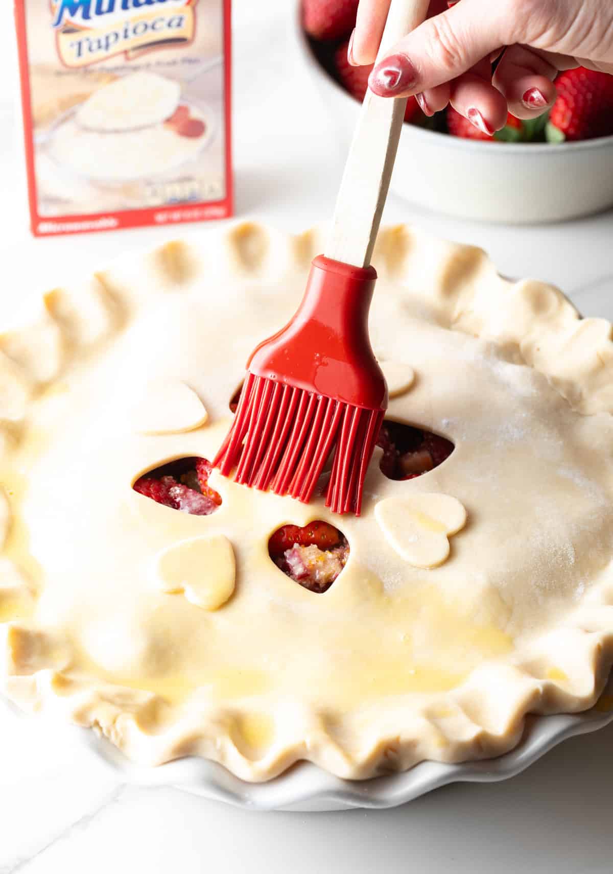 An egg wash is being brushed onto the top of a rhubarb strawberry pie.