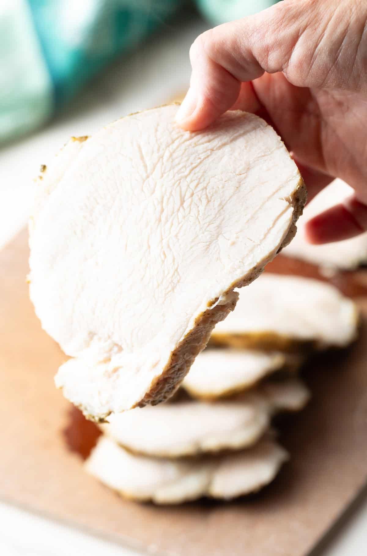 A hand is holding a slice of crock pot turkey breast to the camera above the remainder of the breast.
