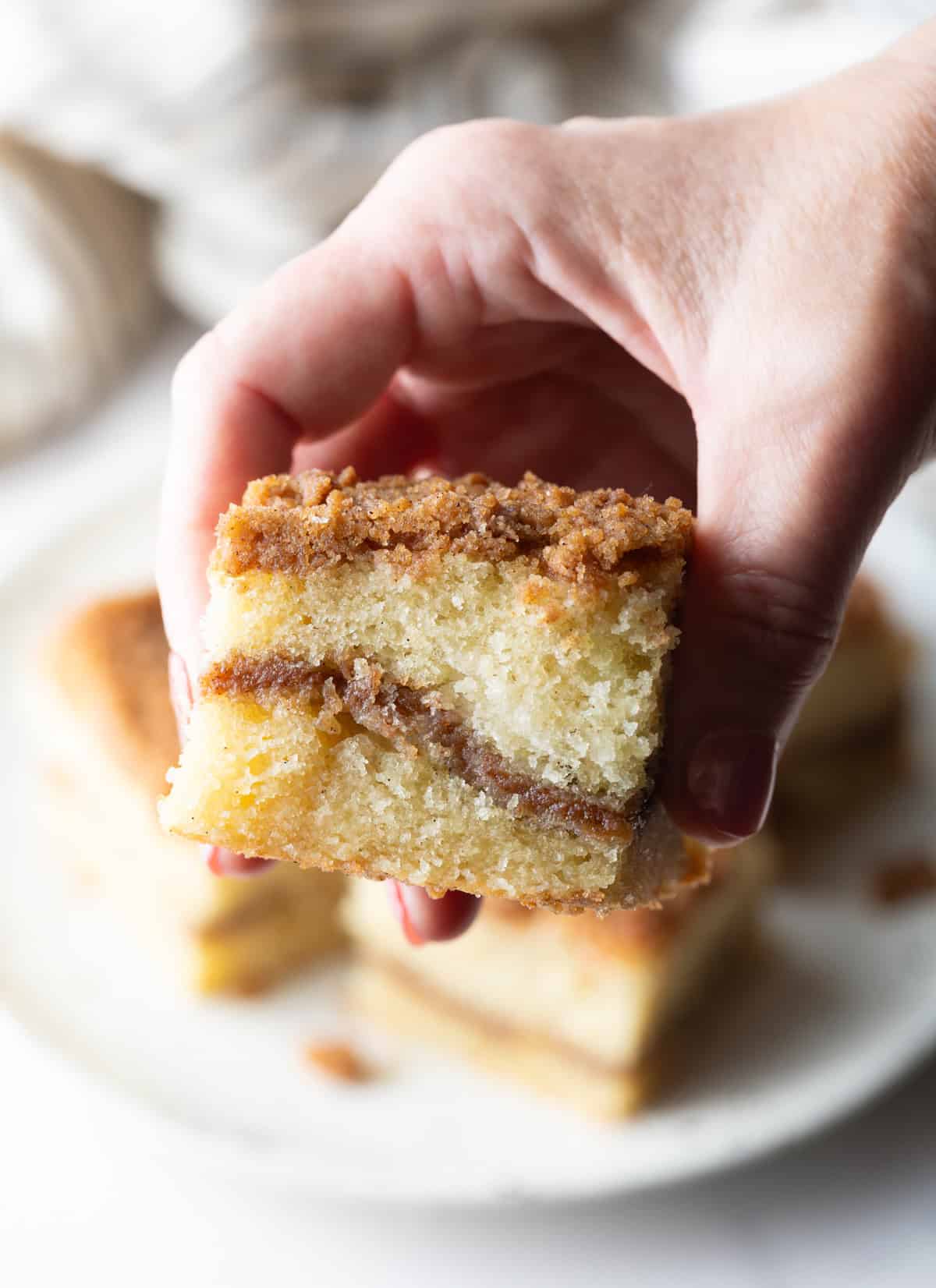 A hand is holding a slice of sour cream coffee cake up to the camera, above a plate of the slices of cake.
