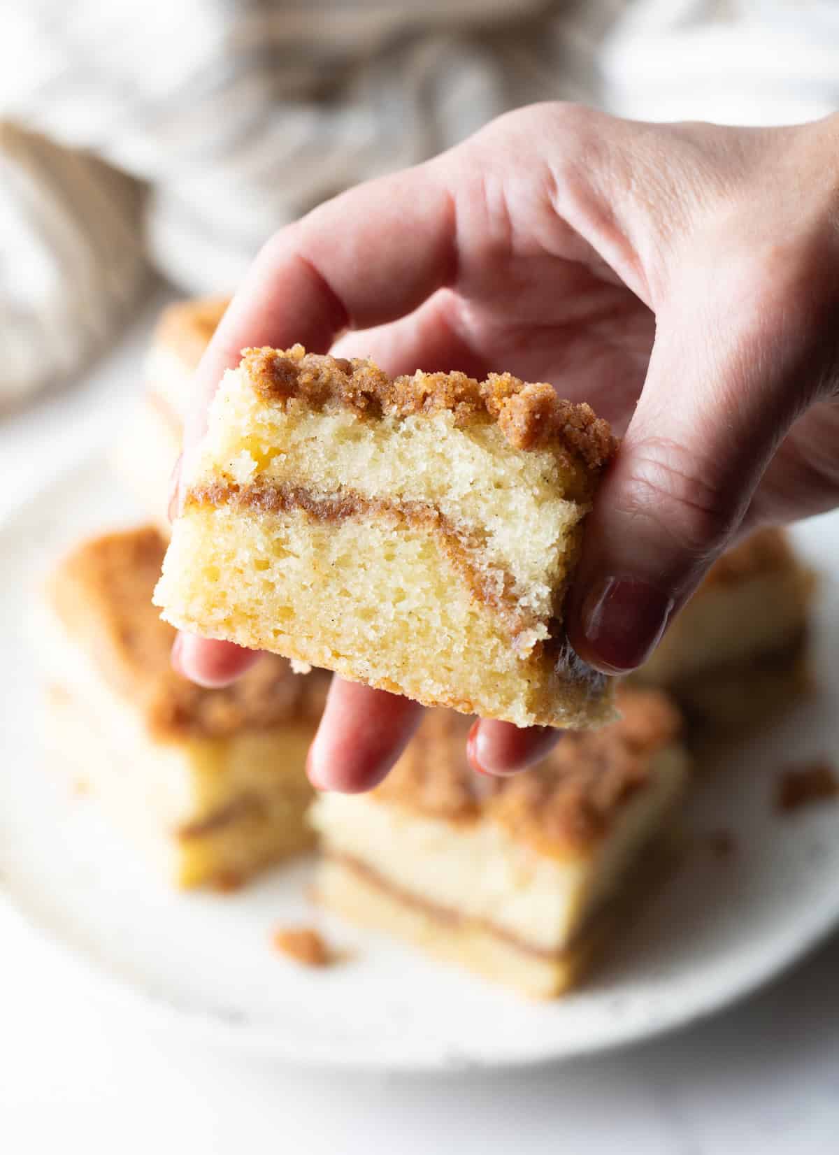 A hand is holding a slice of sour cream coffee cake up to the camera, above a plate of slices of cake.