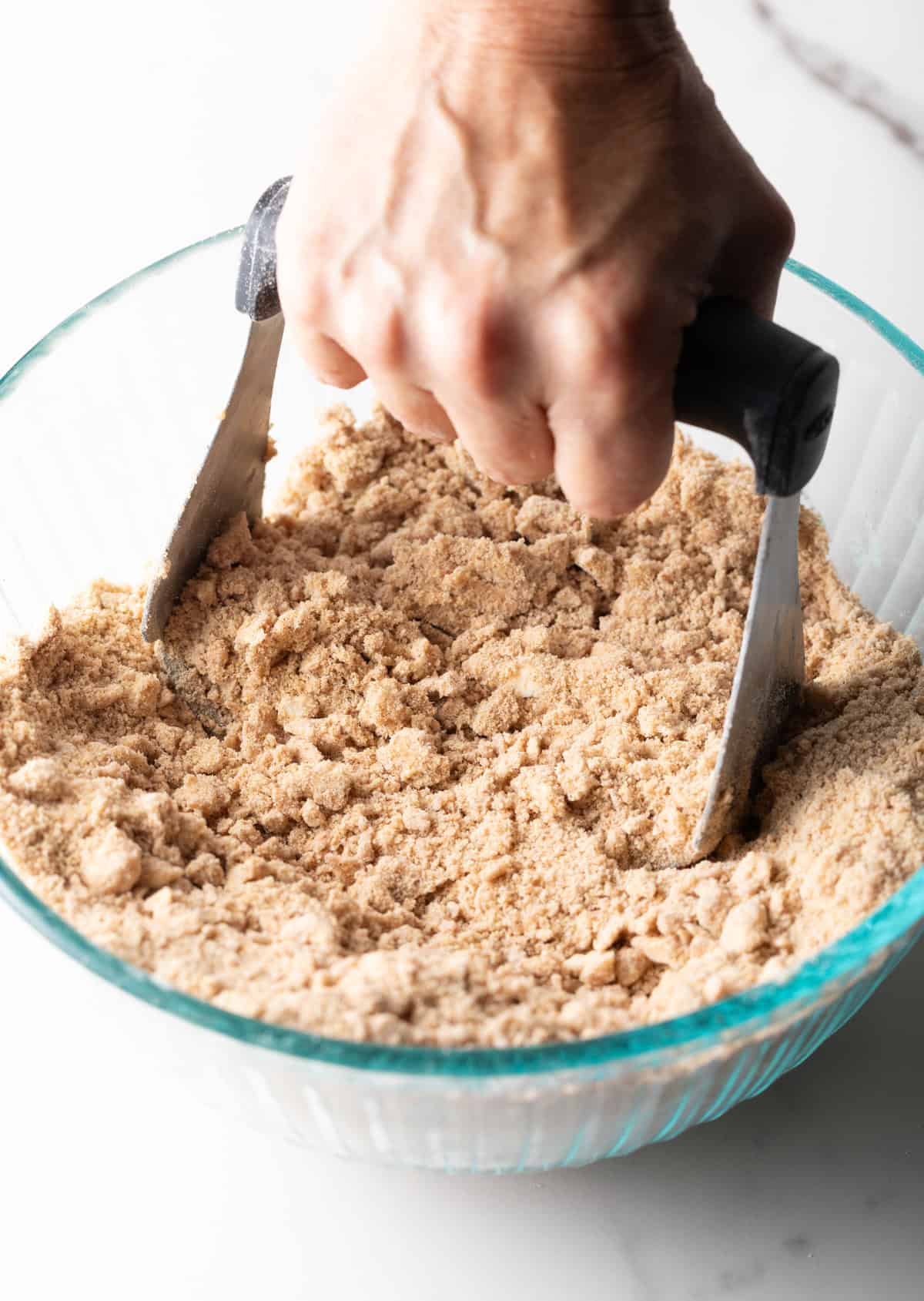 A hand is holding a pastry cutter above a bowl filled with a sour cream coffee cake crumble mixture.
