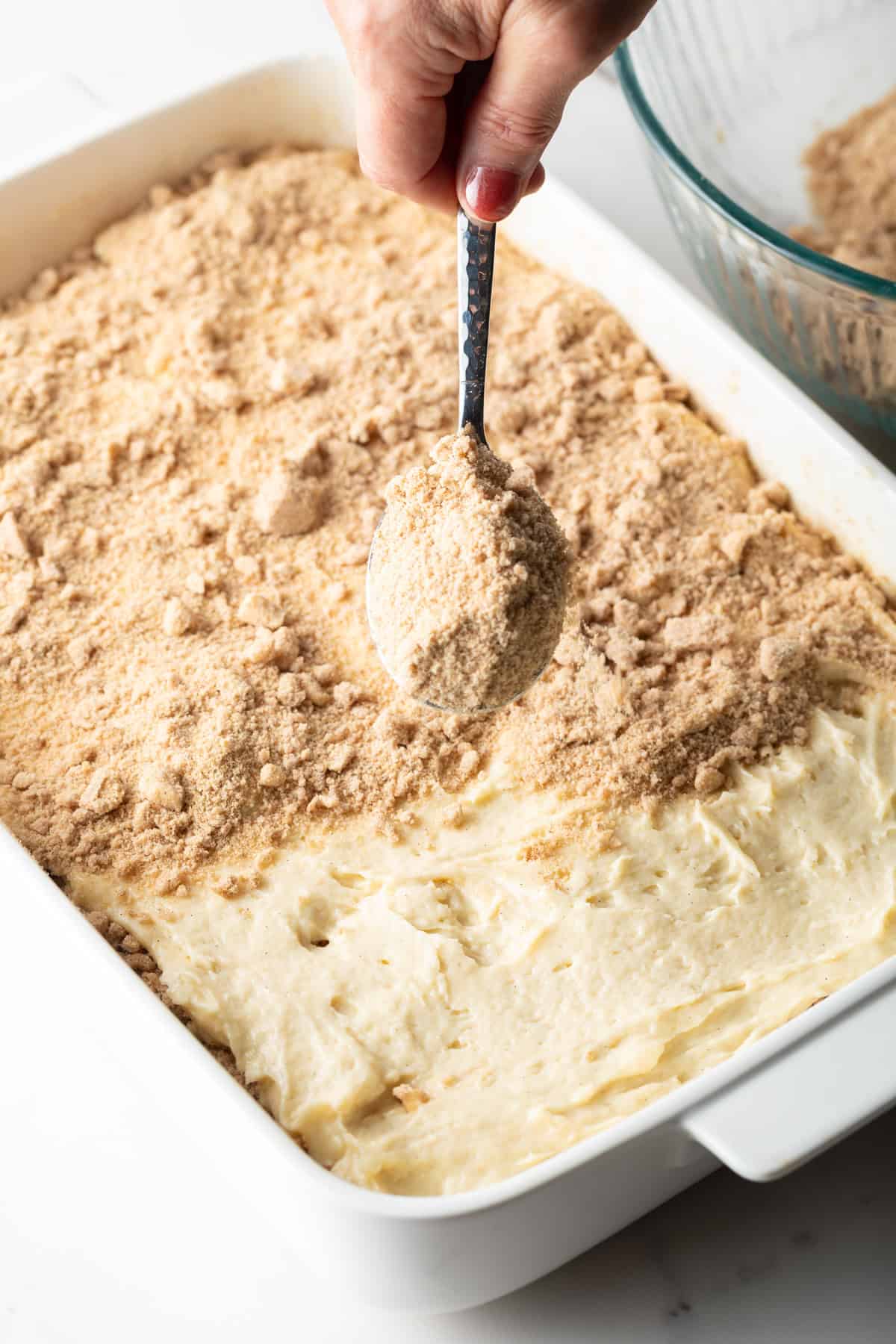 A cinnamon crumb mixture is being added on top of a crumb coffee cake batter with a spoon.