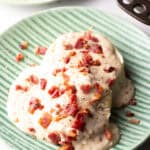 Two biscuits on a green plate, smothered with a bacon gravy recipe, with another plate of biscuits in the background.