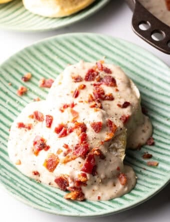 Two biscuits on a green plate, smothered with a bacon gravy recipe, with another plate of biscuits in the background.
