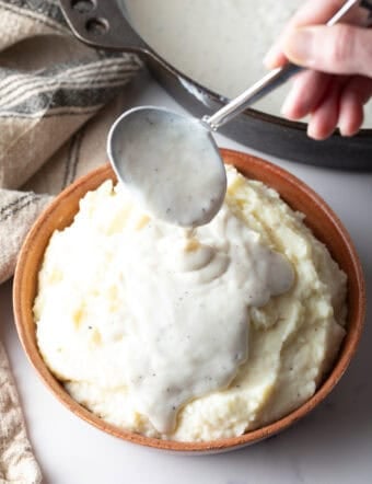 A white gravy recipe is being spooned over a bowl of mashed potatoes.