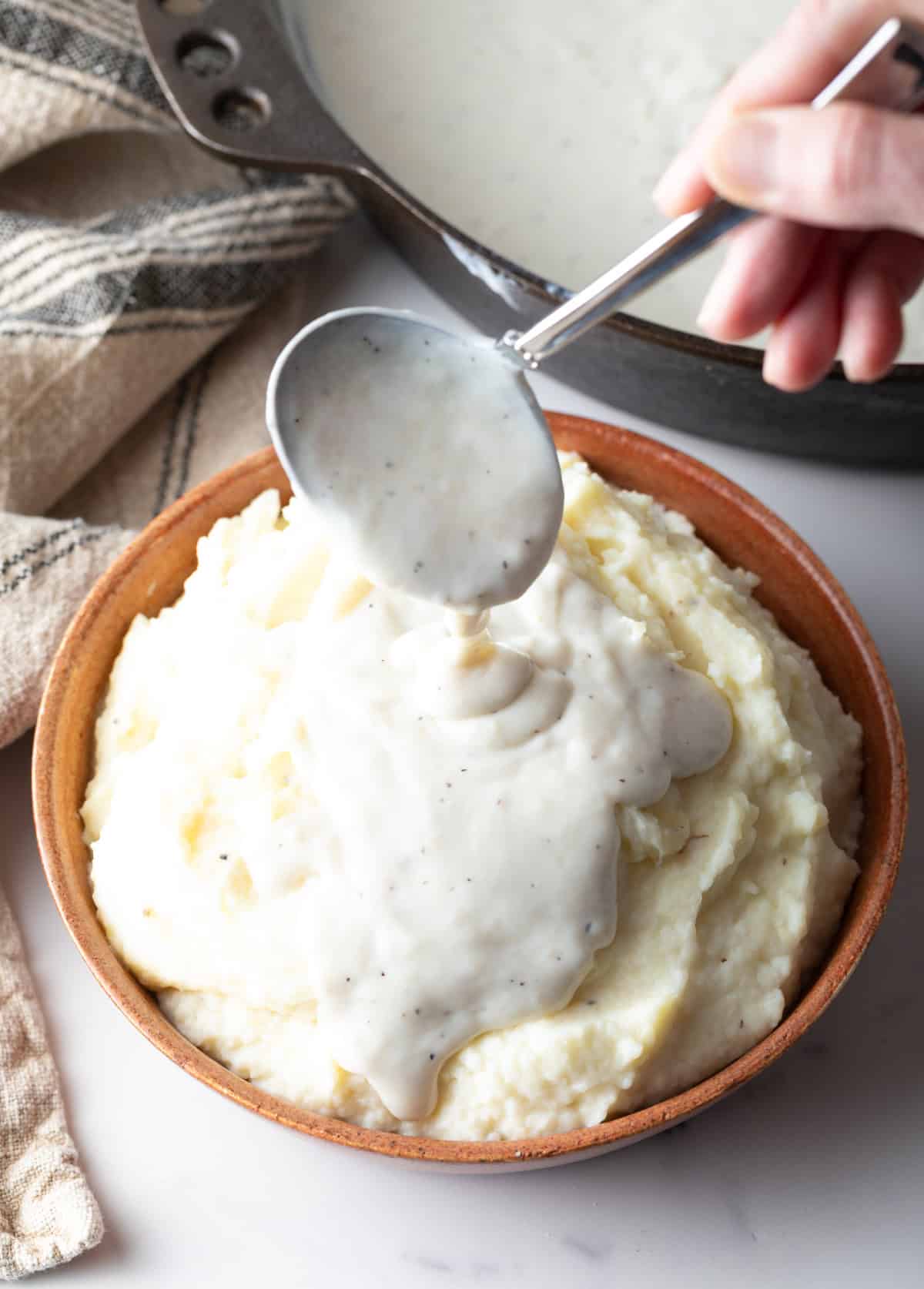 A white gravy recipe is being spooned over a bowl of mashed potatoes.